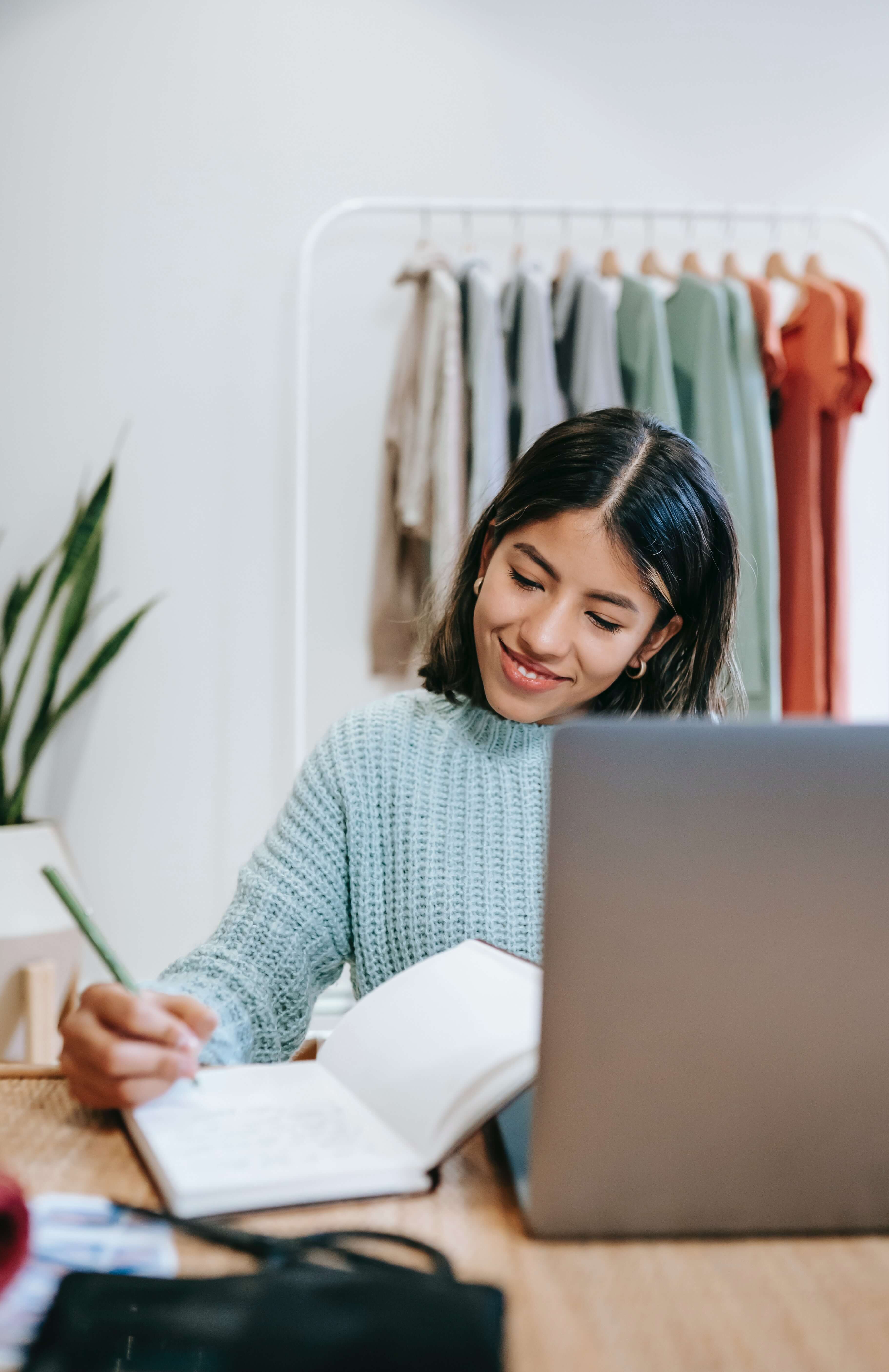 Young woman taking notes at a laptop with clothing rack in the background.